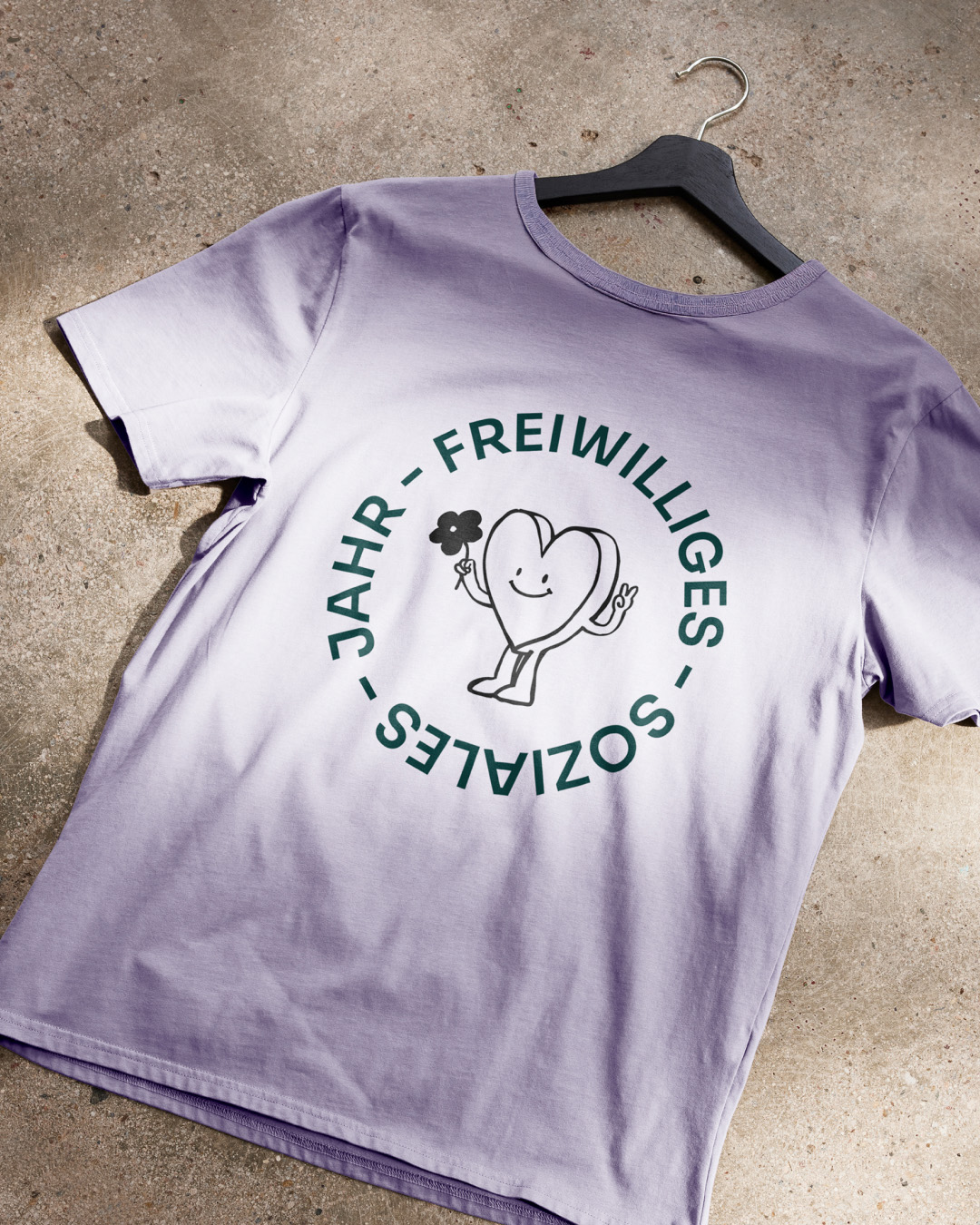Lavender-colored T-shirt on a hanger featuring the circular text “Freiwilliges Soziales Jahr” and a friendly heart illustration – part of the FSJ branding designed by Vienna-based creative agency Achtzehn Grad.