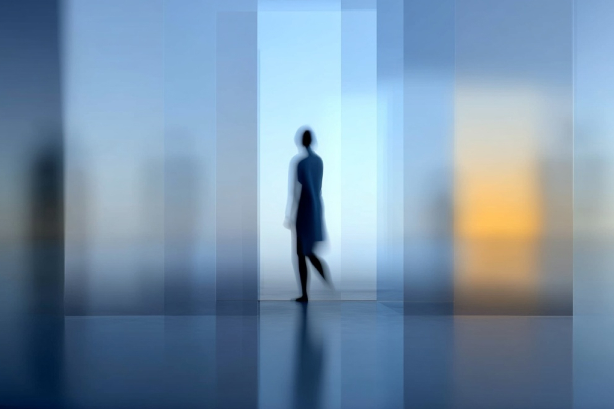 Unscharfe Silhouette einer gehenden Person zwischen transparenten Glasflächen in kühlen Blau- und warmen Lichttönen.
