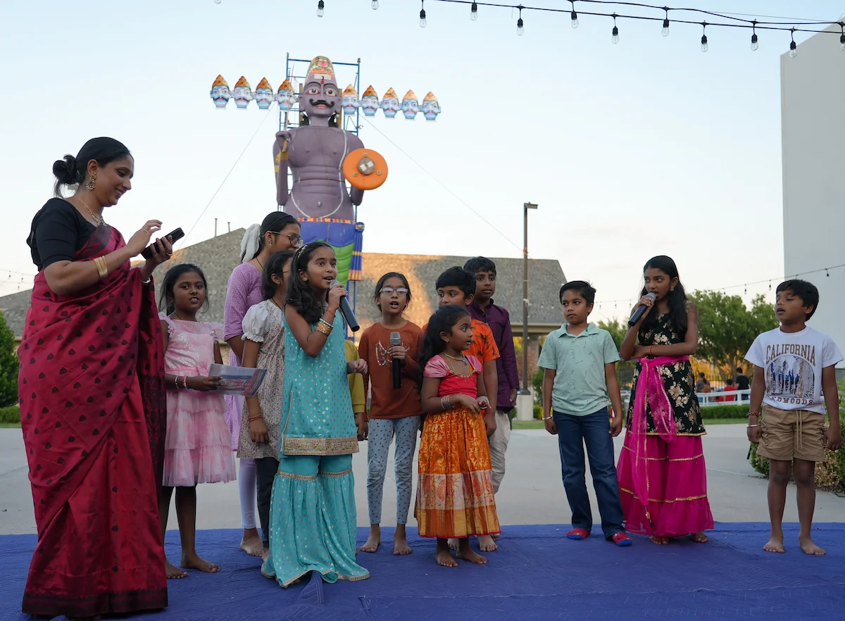 Children singing on an outdoor stage with the Ravan effigy behind them.