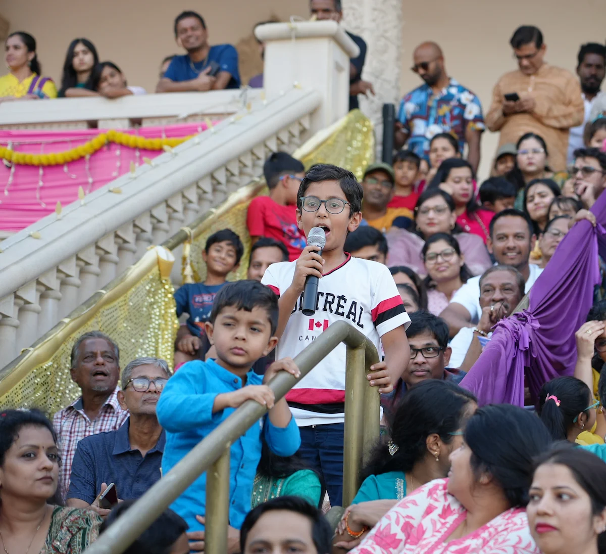 A young participant speaking into a microphone among families in the temple courtyard.