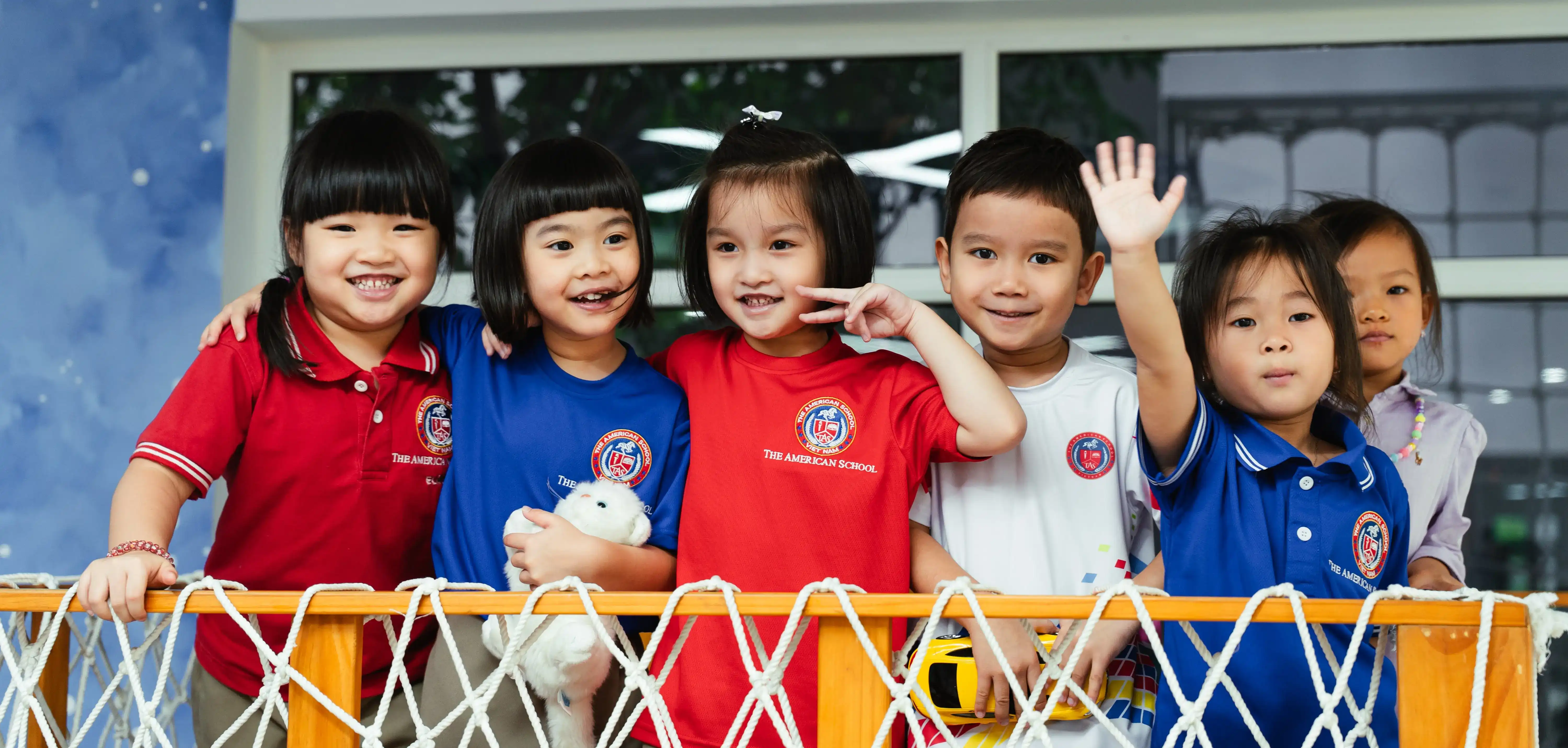 Five smiling children wearing colorful school uniforms, standing behind a wooden railing with white rope netting, one child waving and another holding a toy car.