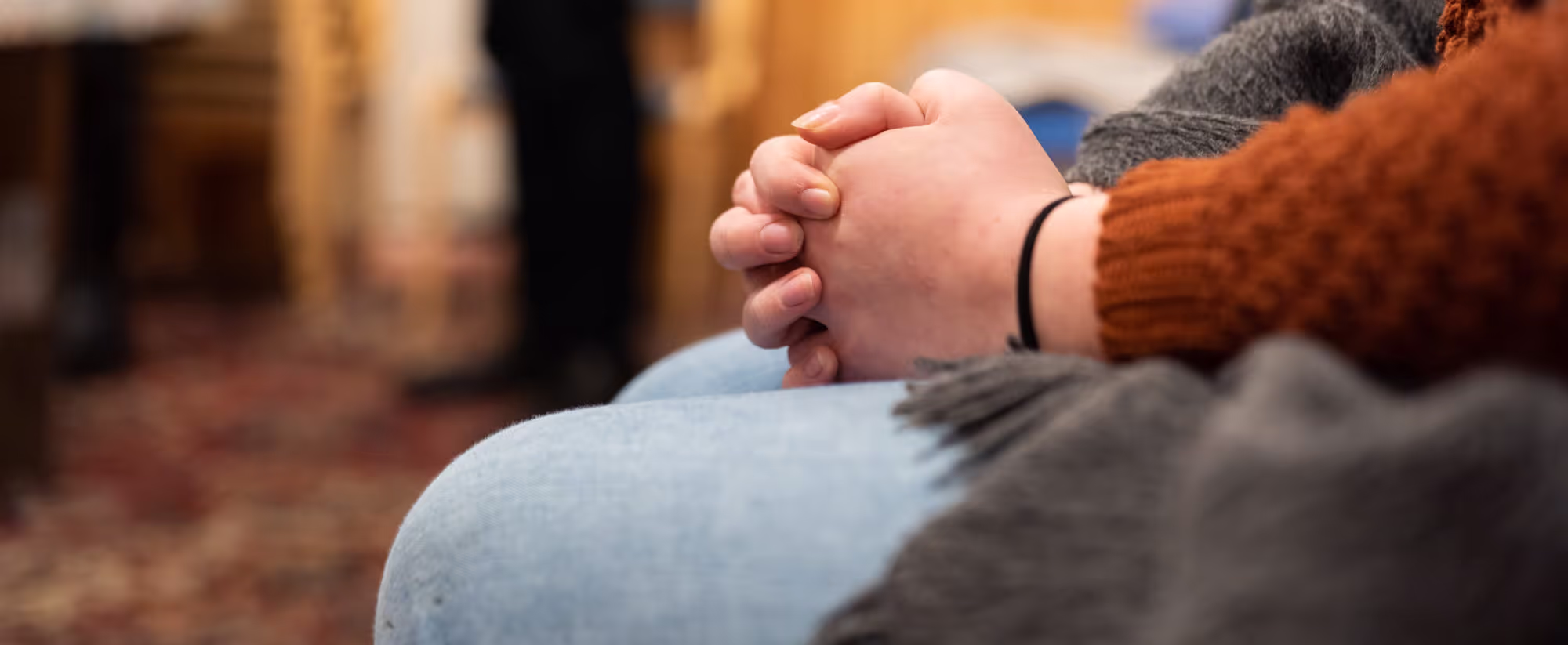 Nærbilde av to hender som er foldet sammen, plassert på fanget til en person iført lyse blå jeans og en oransje genser.