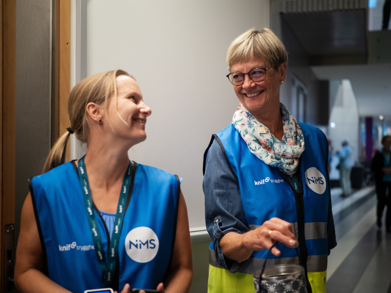 To kvinner iført blå sikkerhetsvester med NIMS-logo som smiler og ser på hverandre i en korridor.