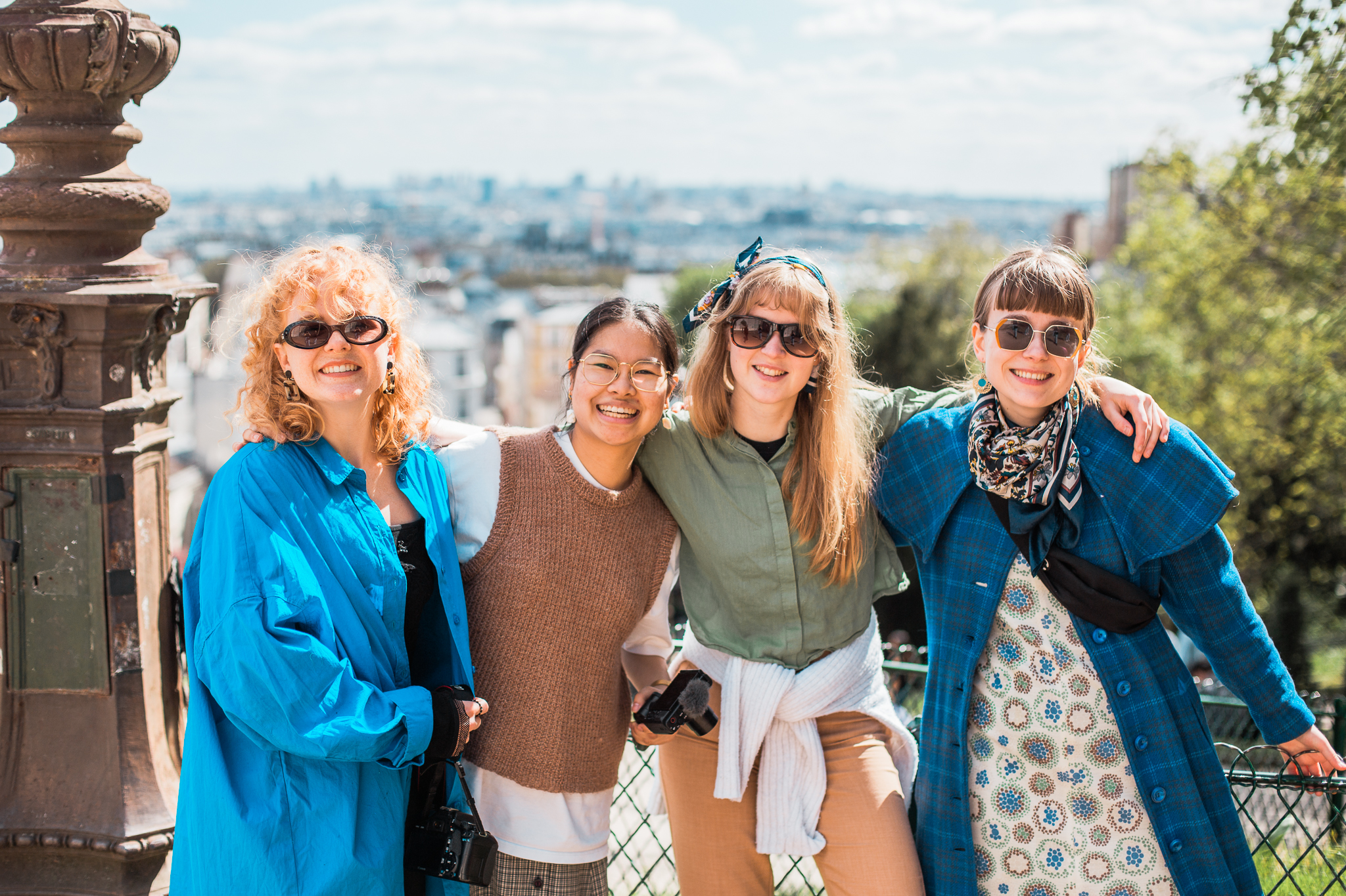 Fire kvinner smilende med solbriller og armene rundt hverandre utendørs mot en bybakgrunn.