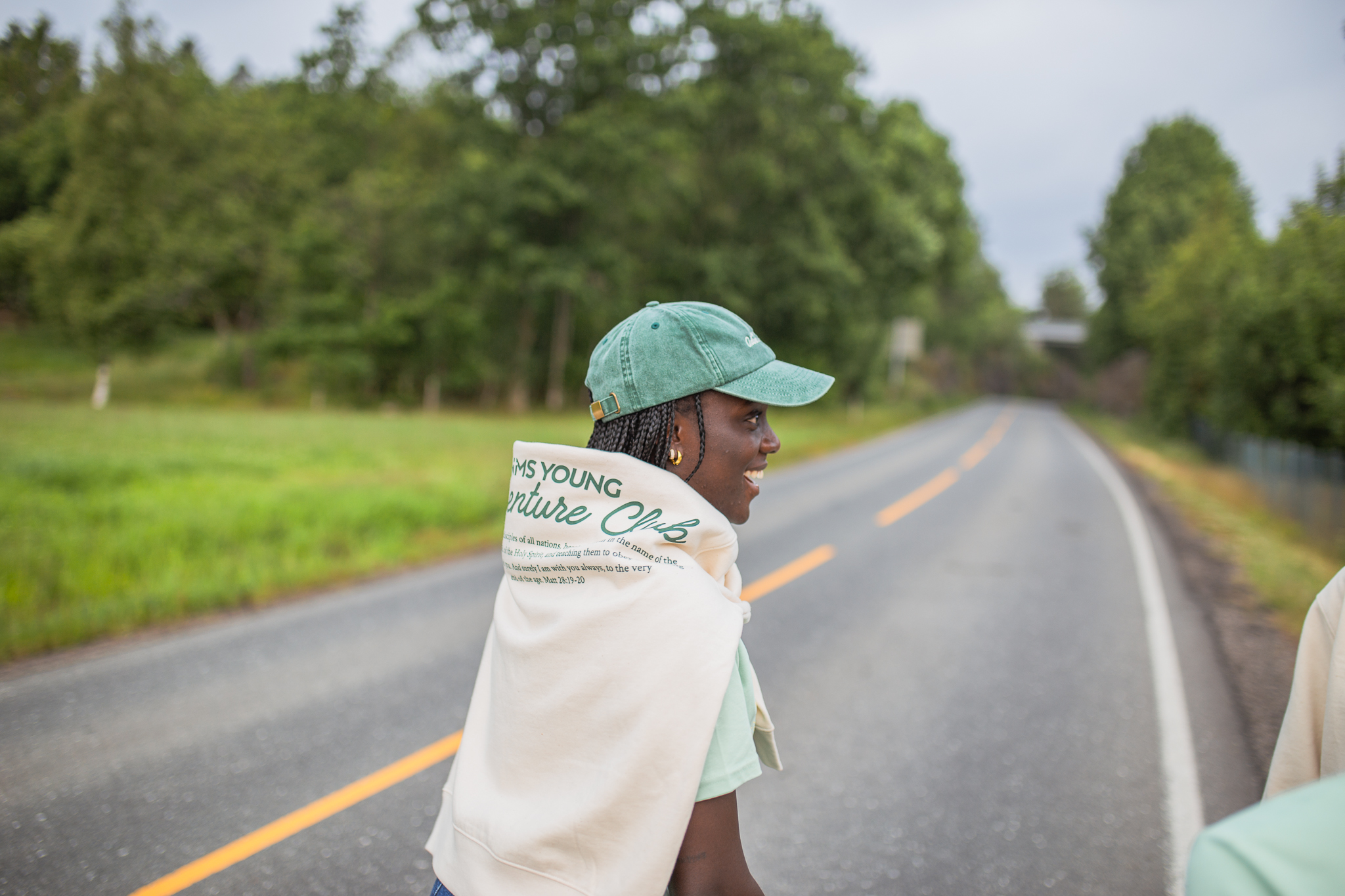 Smilende person med grønn caps og hvit hettegenser kastet over skuldrene står ved en landevei omgitt av trær.