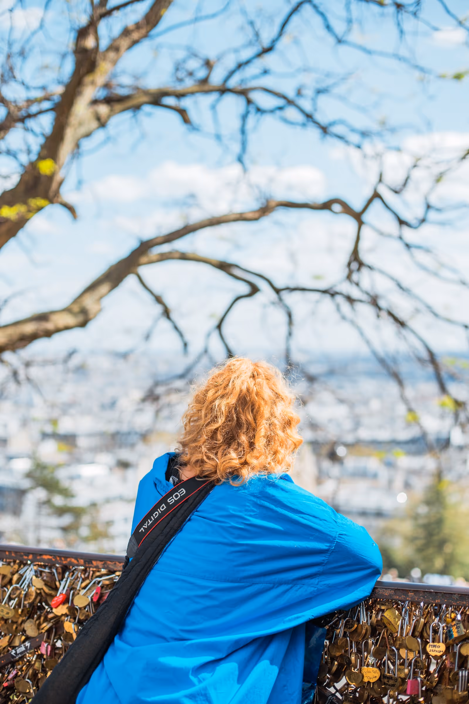 Person med krøllete rødt hår og blå jakke ser utover byen fra et gjerde dekket med hengelåser.