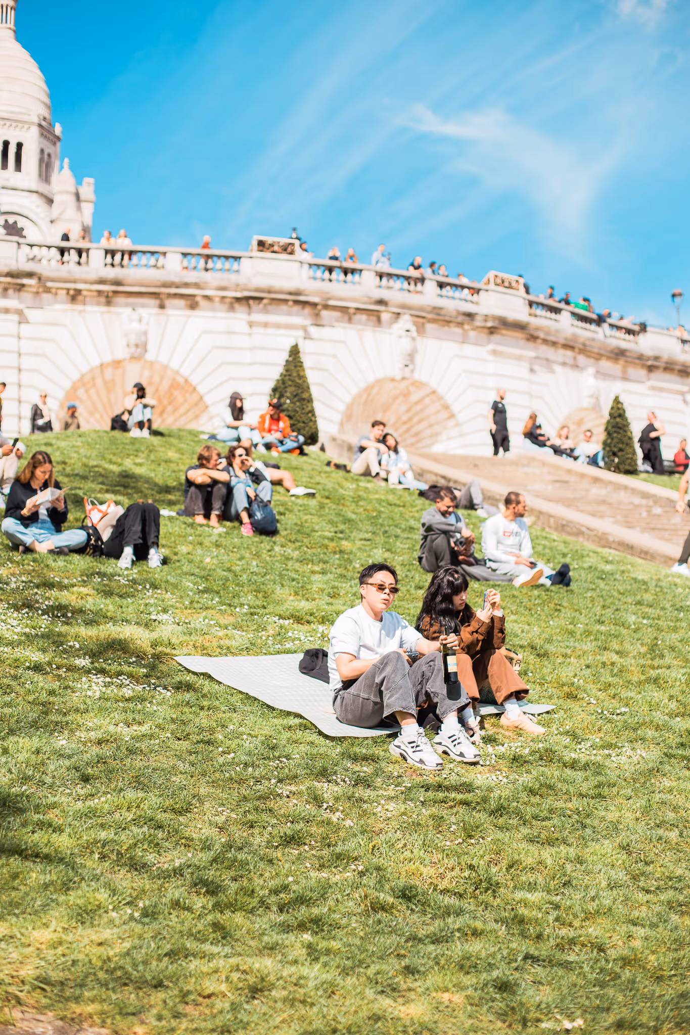 Folk som sitter og slapper av på en solrik gressbakke ved en historisk bygning med trapp og mennesker på toppen av muren.