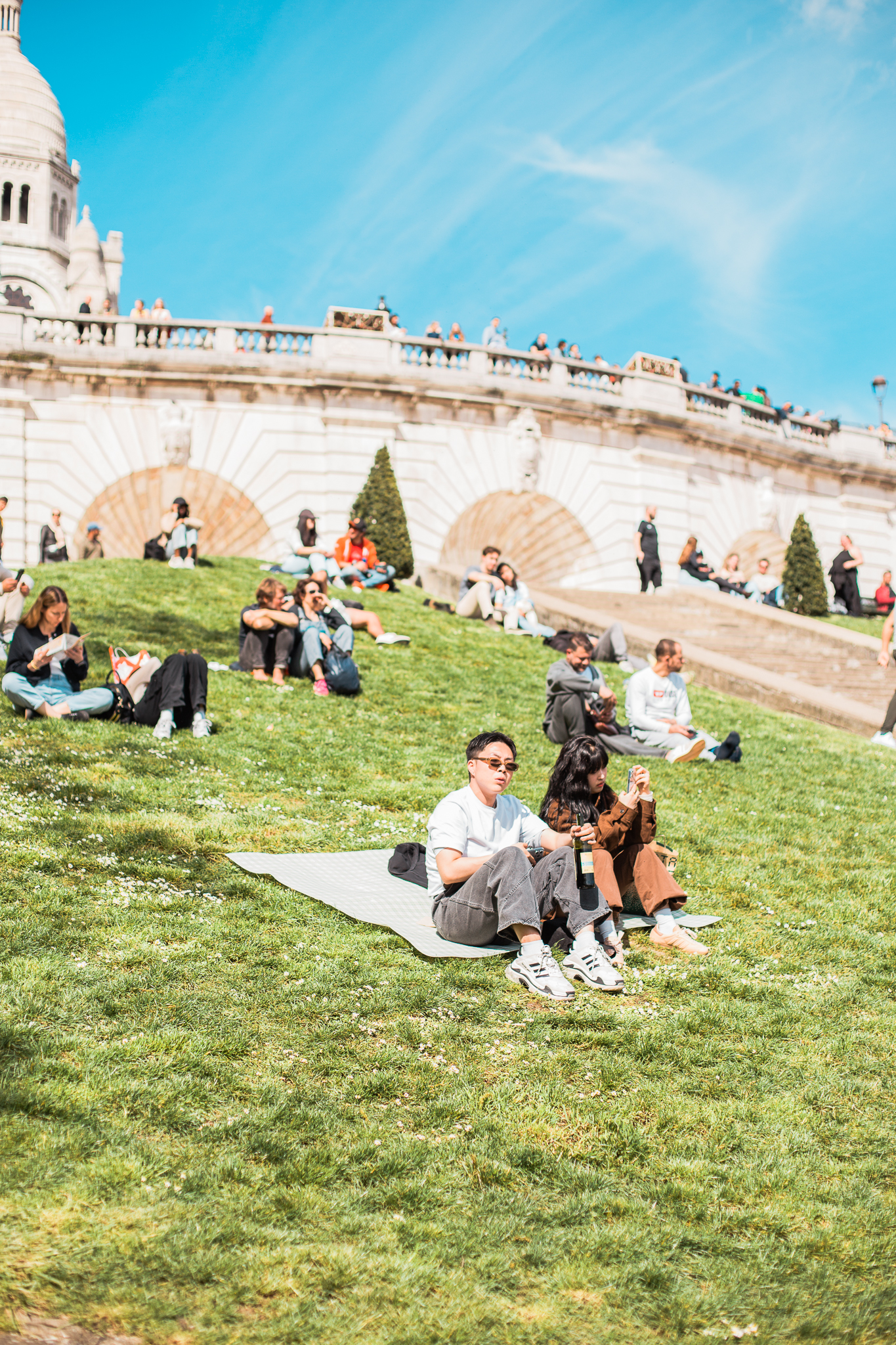 Folk som sitter og slapper av på en solrik gressbakke ved en historisk bygning med trapp og mennesker på toppen av muren.