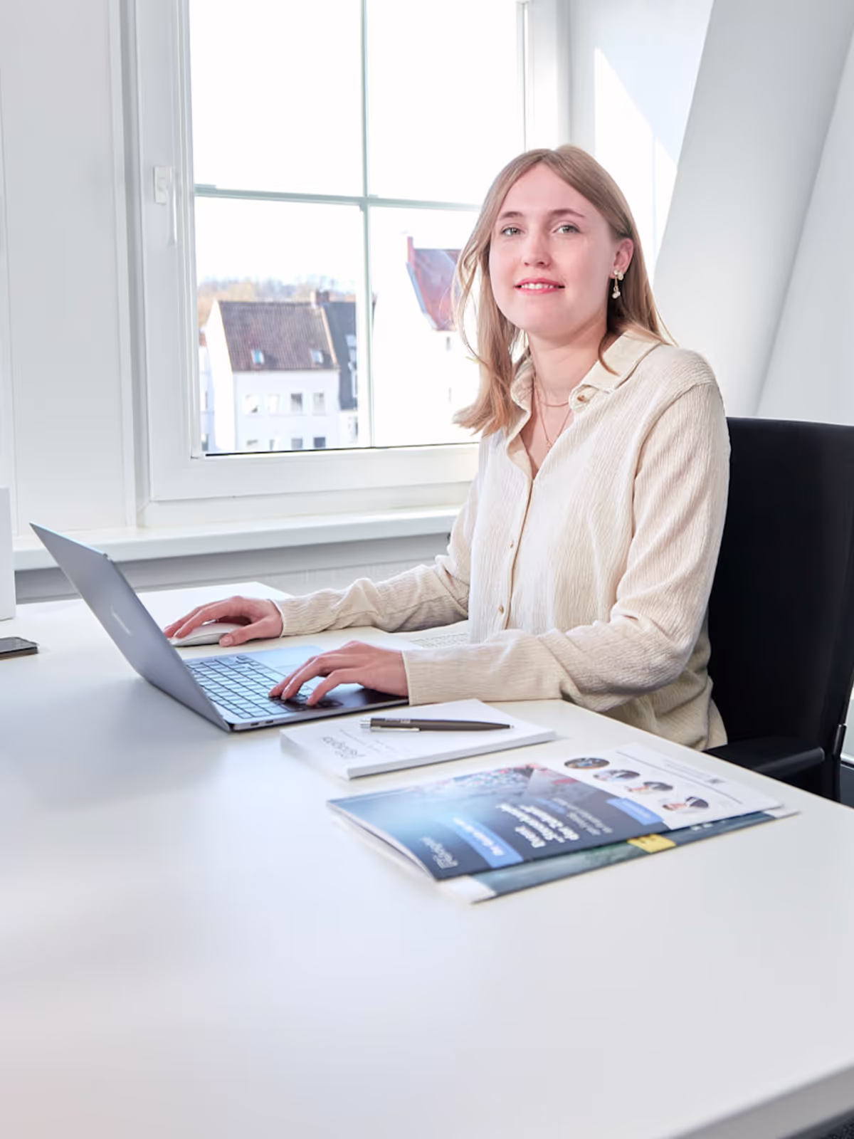 Eine junge Frau sitzt an einem weißen Schreibtisch und arbeitet an einem Laptop in einem hellen Büro mit großem Fenster. Auf dem Tisch liegen Informationsmaterialien und ein Stift, sie blickt freundlich in die Kamera.