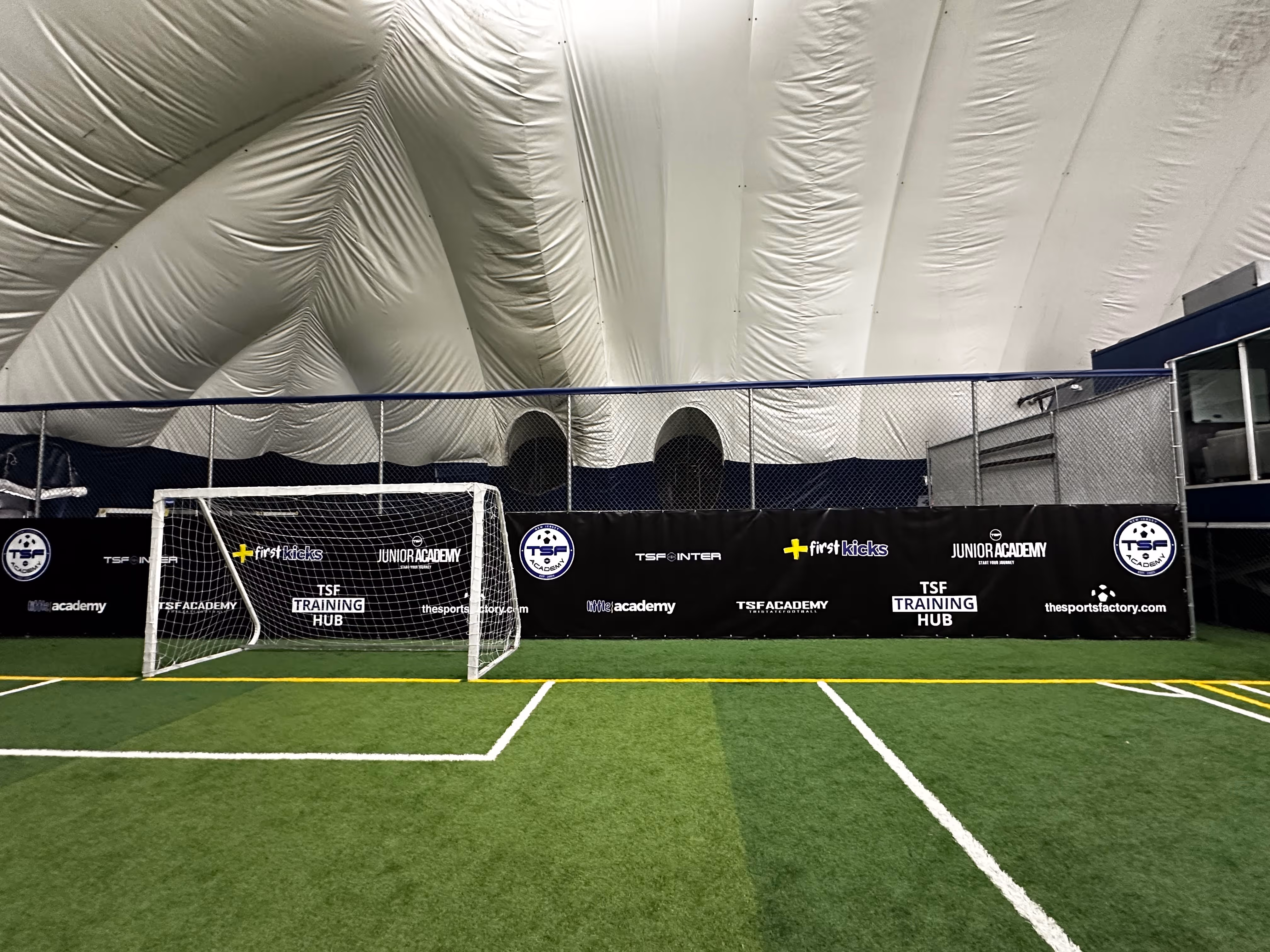 Indoor soccer field with artificial turf, a white goalpost, and a black fence displaying logos for TSF Training Hub and Junior Academy.