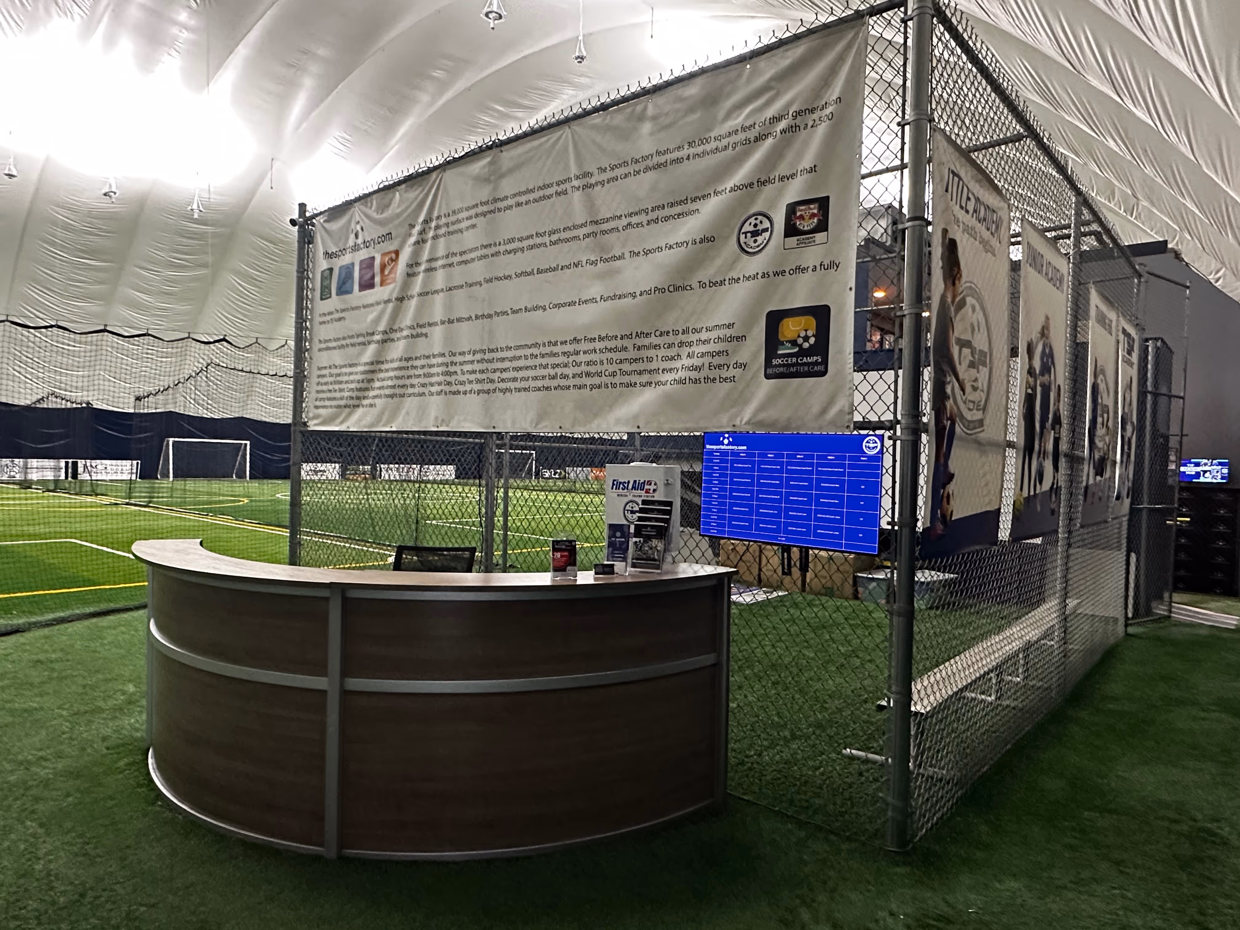 Reception desk inside a large indoor sports facility with artificial turf, soccer goals, chain-link fencing, informational banners, and digital monitors.