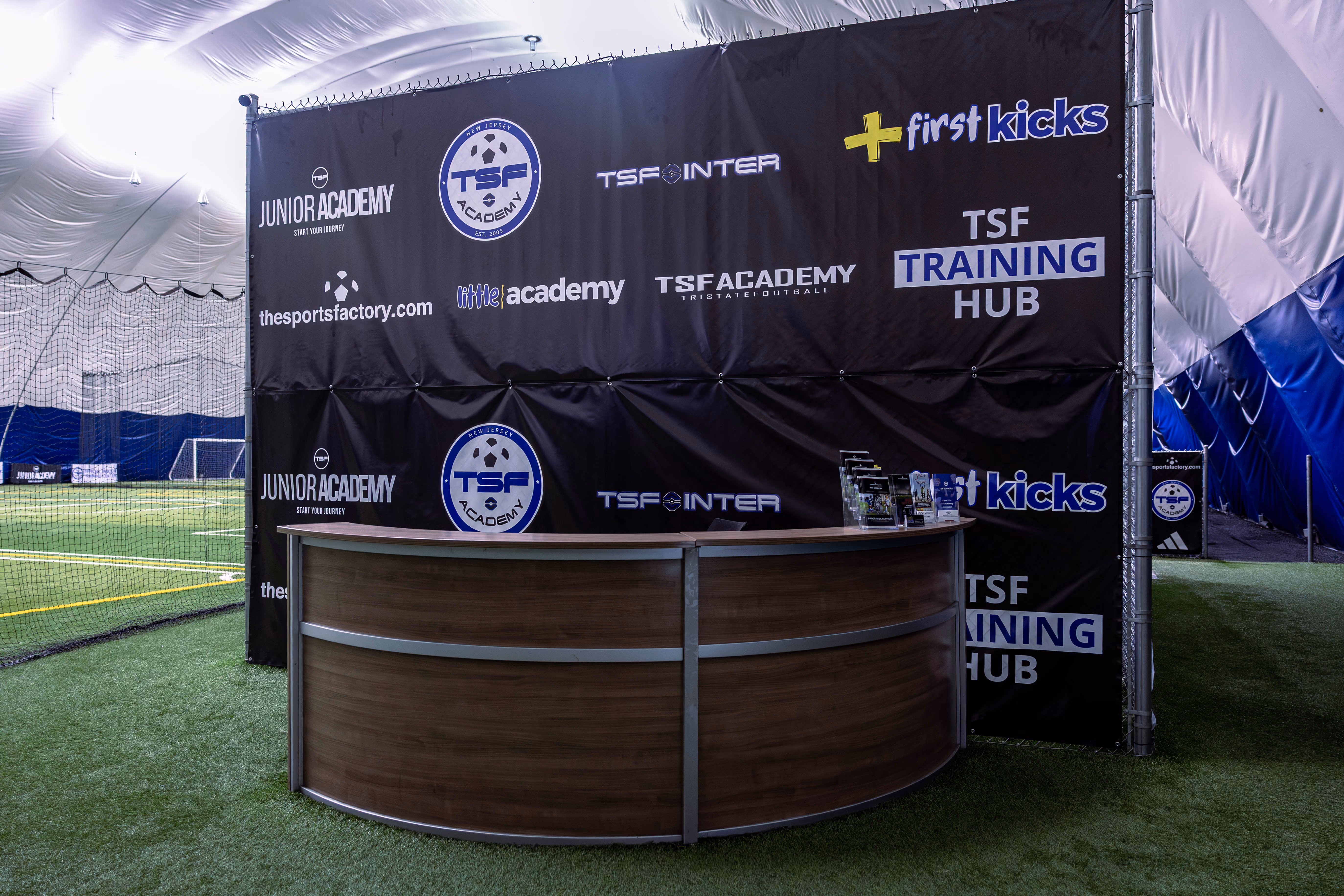 Indoor sports facility with artificial turf featuring a curved wooden reception desk in front of a black backdrop displaying logos for TSF Academy, Junior Academy, TSF Inter, First Kicks, and TSF Training Hub.