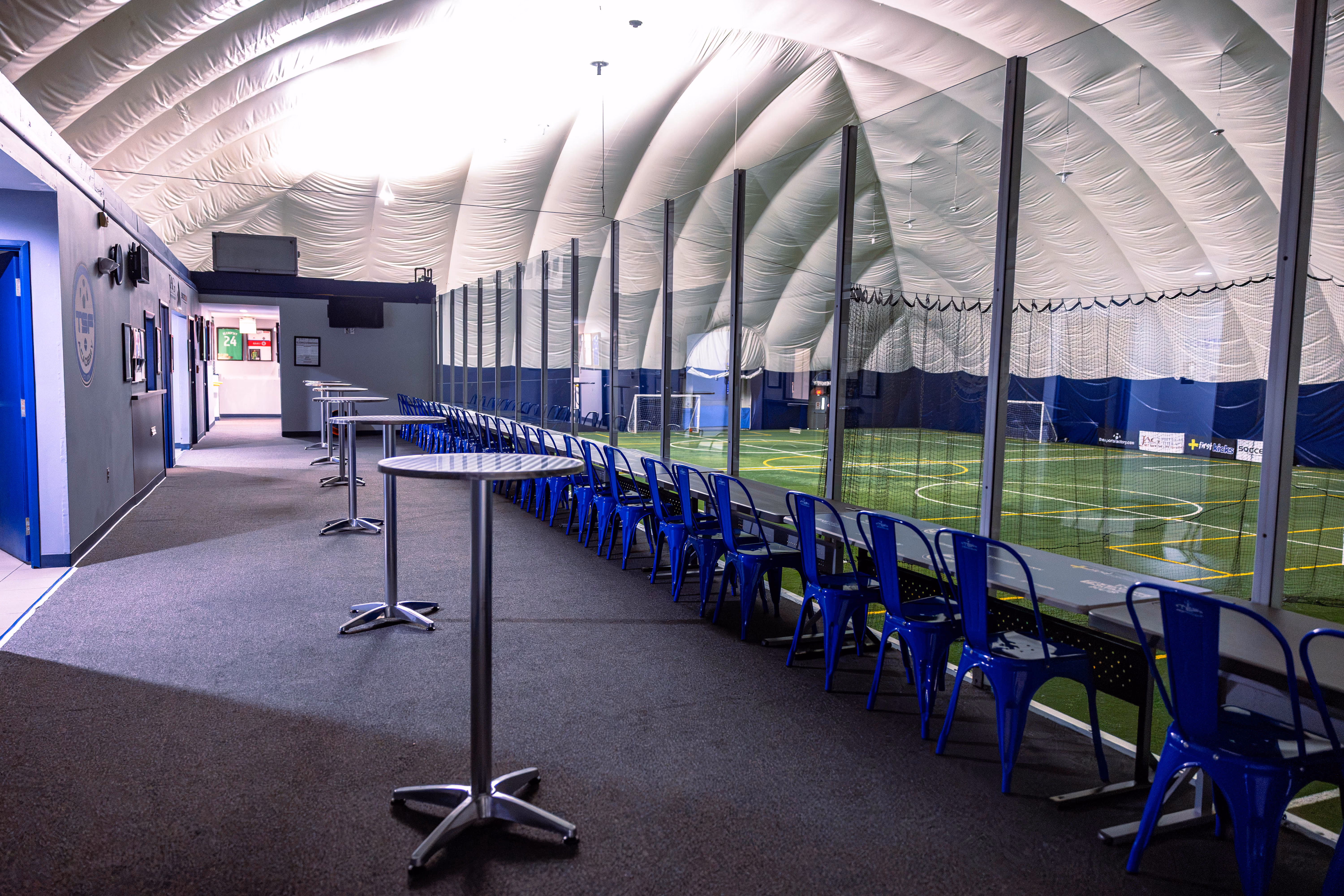 Interior of a sports facility with a row of blue chairs and tables along a glass partition overlooking an indoor turf field inside a large white dome structure.