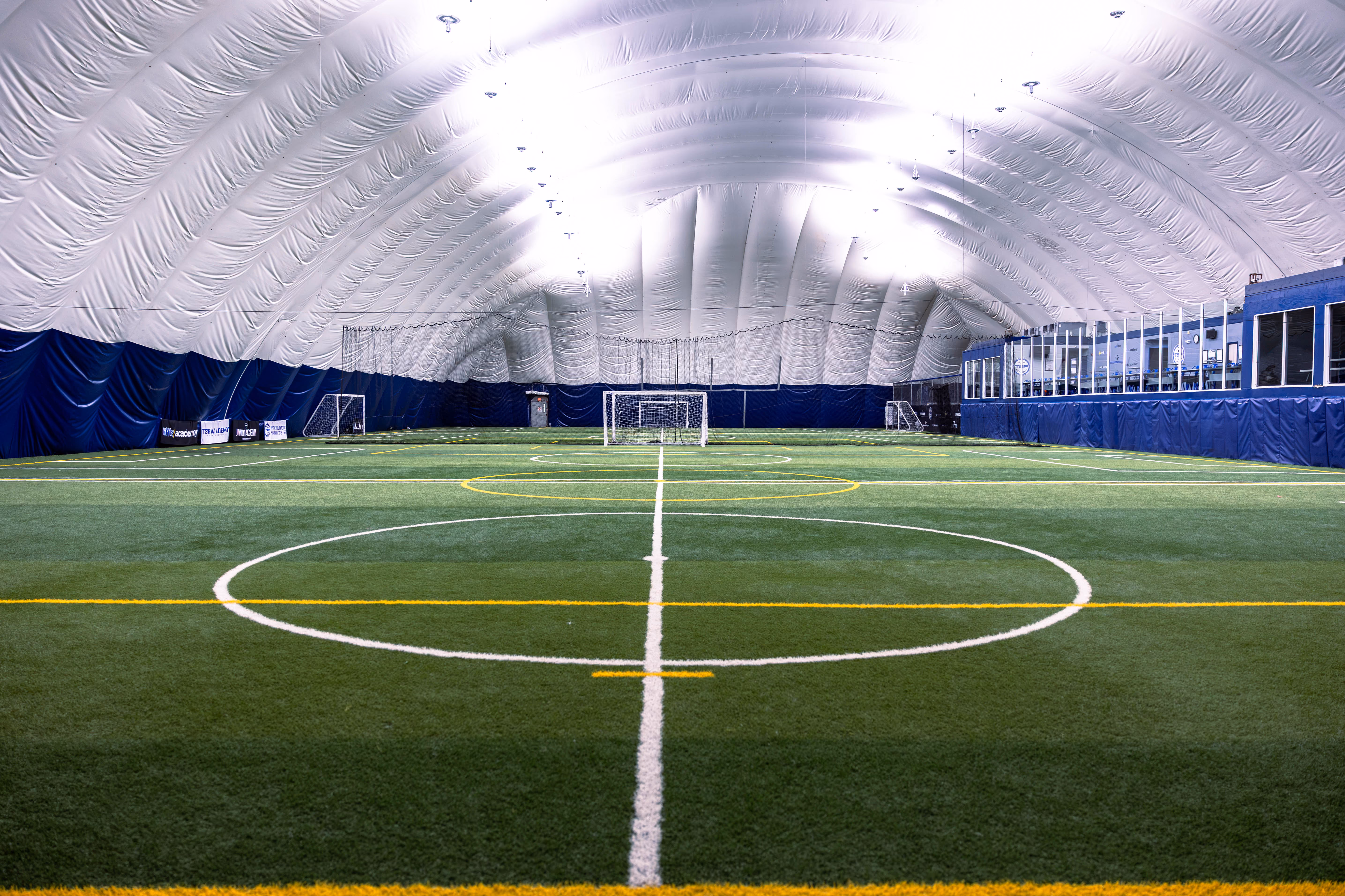 Indoor soccer field with artificial turf inside a large white inflatable dome, featuring white and yellow field markings and multiple soccer goals along the walls.