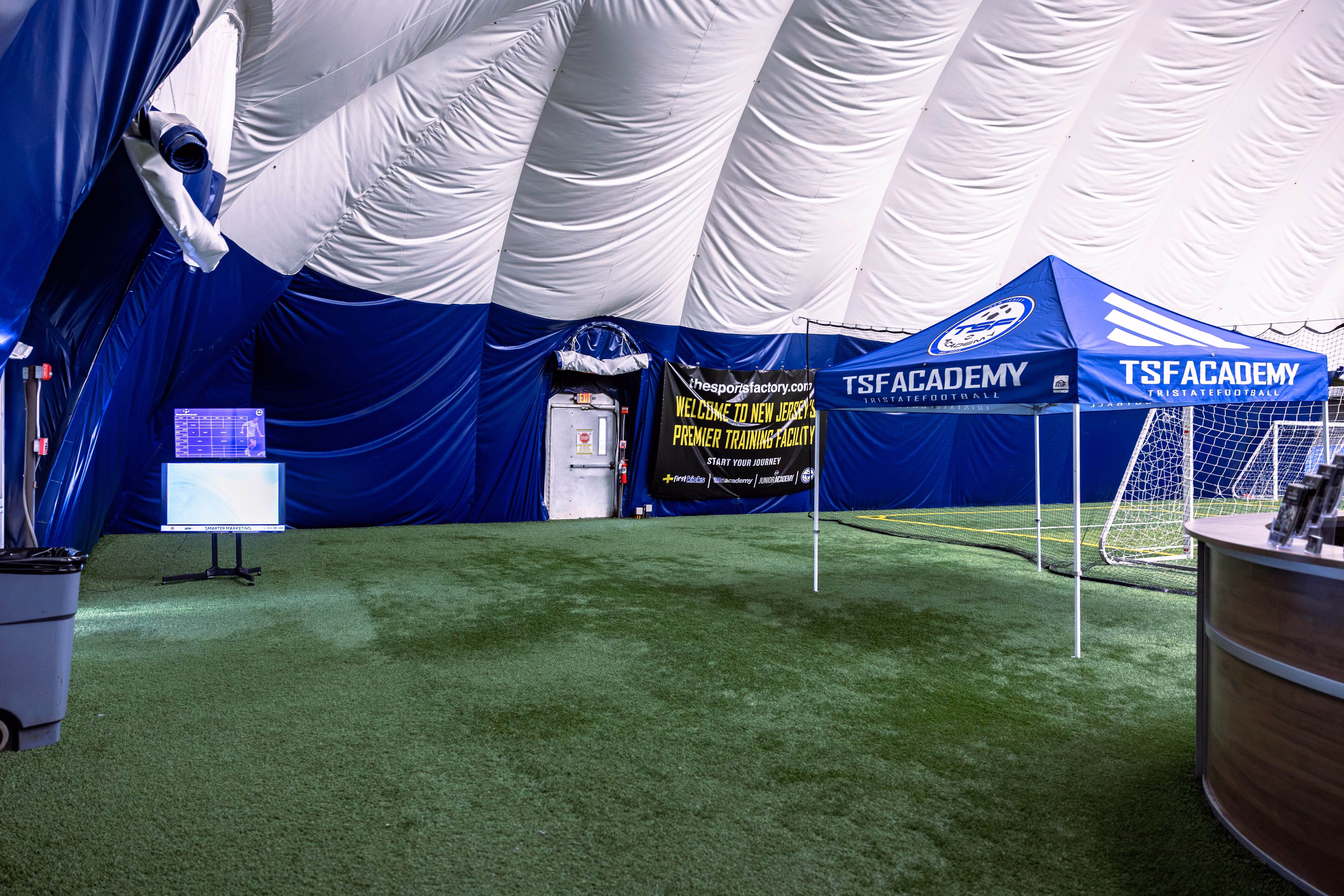 Indoor sports facility with artificial turf, a blue and white TSF Academy tent, soccer goals, and a banner welcoming to New Jersey's premier training facility.