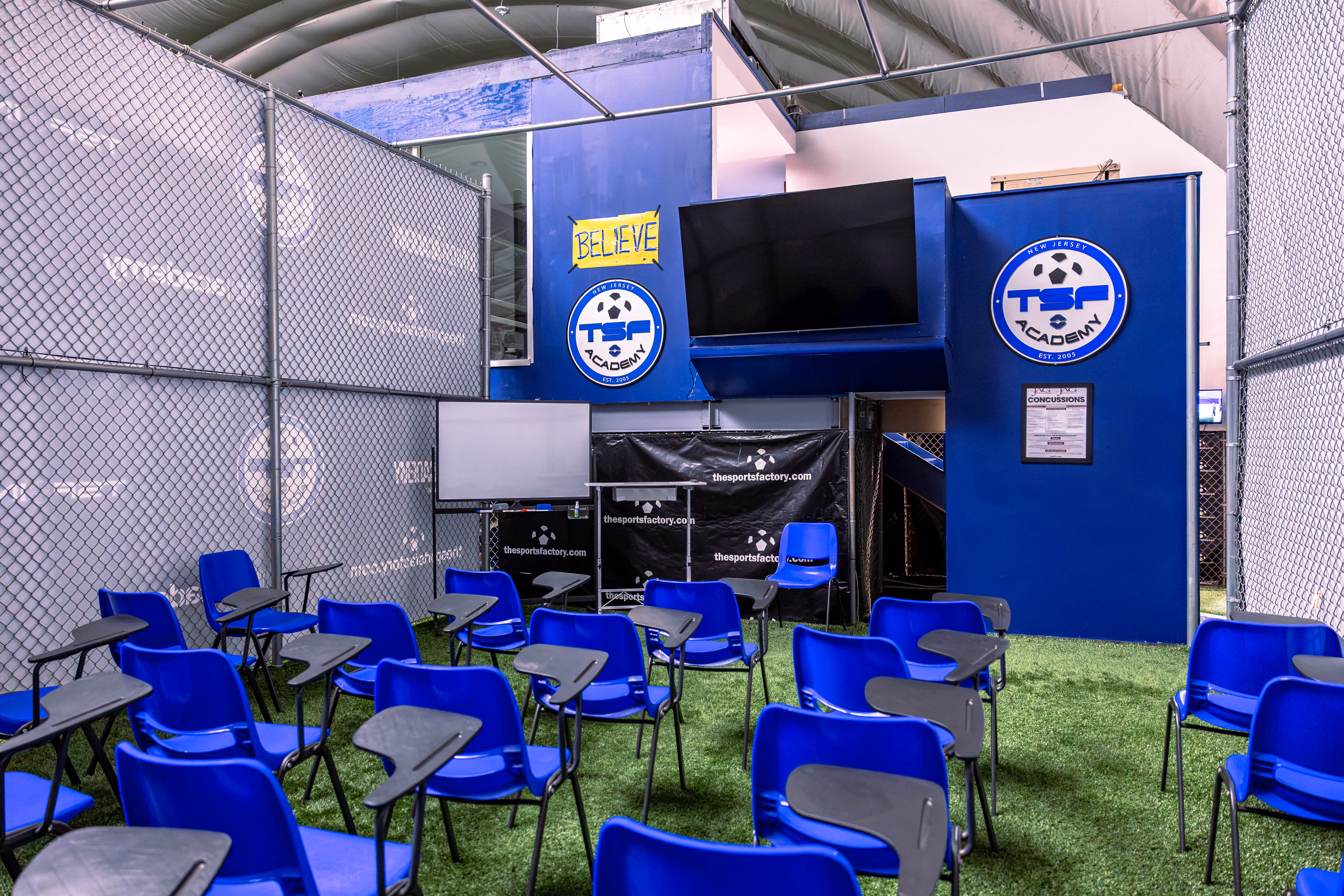 Indoor classroom with blue plastic chairs and attached small desks arranged on artificial turf, surrounded by chain-link fencing, featuring a large flat-screen TV mounted on a blue wall with TSF Academy logos and a yellow sign that says Believe.
