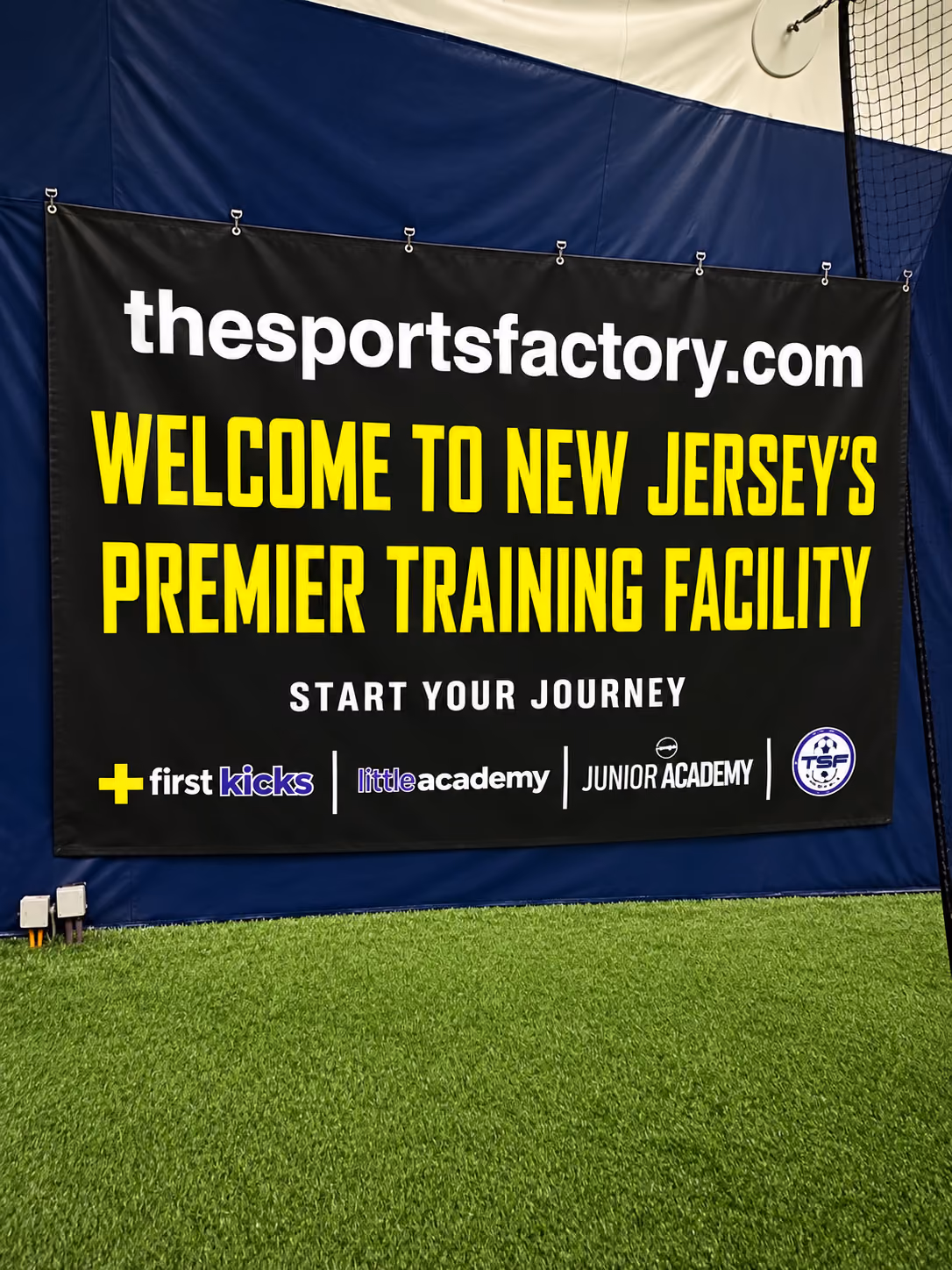 Indoor sports training facility with a large black banner reading 'thesportsfactory.com, Welcome to New Jersey's Premier Training Facility, Start Your Journey' above logos for First Kicks, Little Academy, and Junior Academy on artificial green turf.