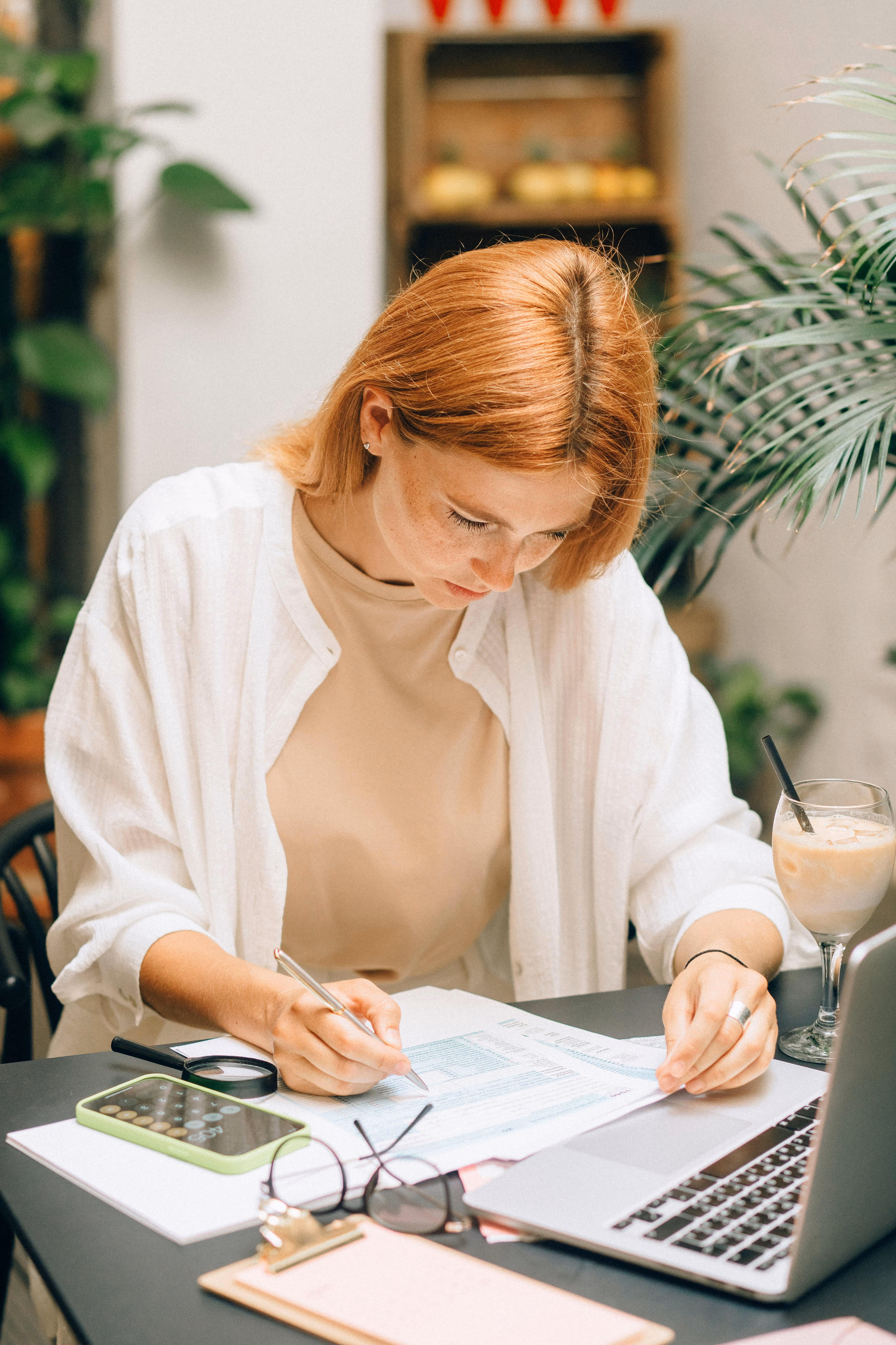 Frau mit roten Haaren arbeitet an Dokumenten am Tisch mit Laptop, Smartphone und Getränk.