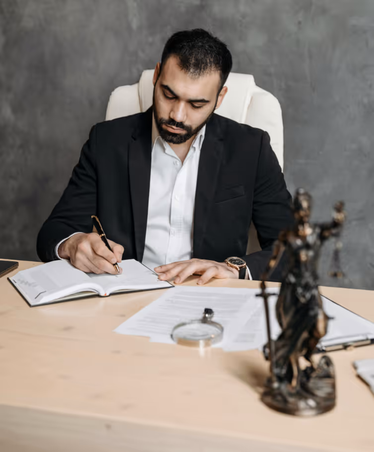 Ein Mann mit Bart und dunklem Anzug, der in einem weißen Stuhl sitzt und in einem Buch auf einem Schreibtisch schreibt.