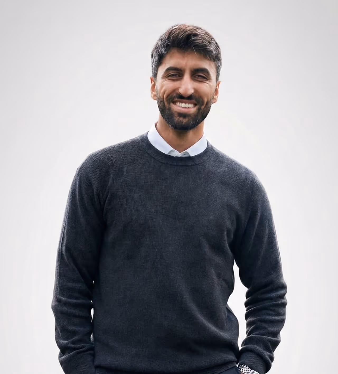 Lächelnder Mann mit dunklem Bart und kurzen Haaren, trägt dunkelgrauen Pullover über weißem Hemd vor hellem Hintergrund.