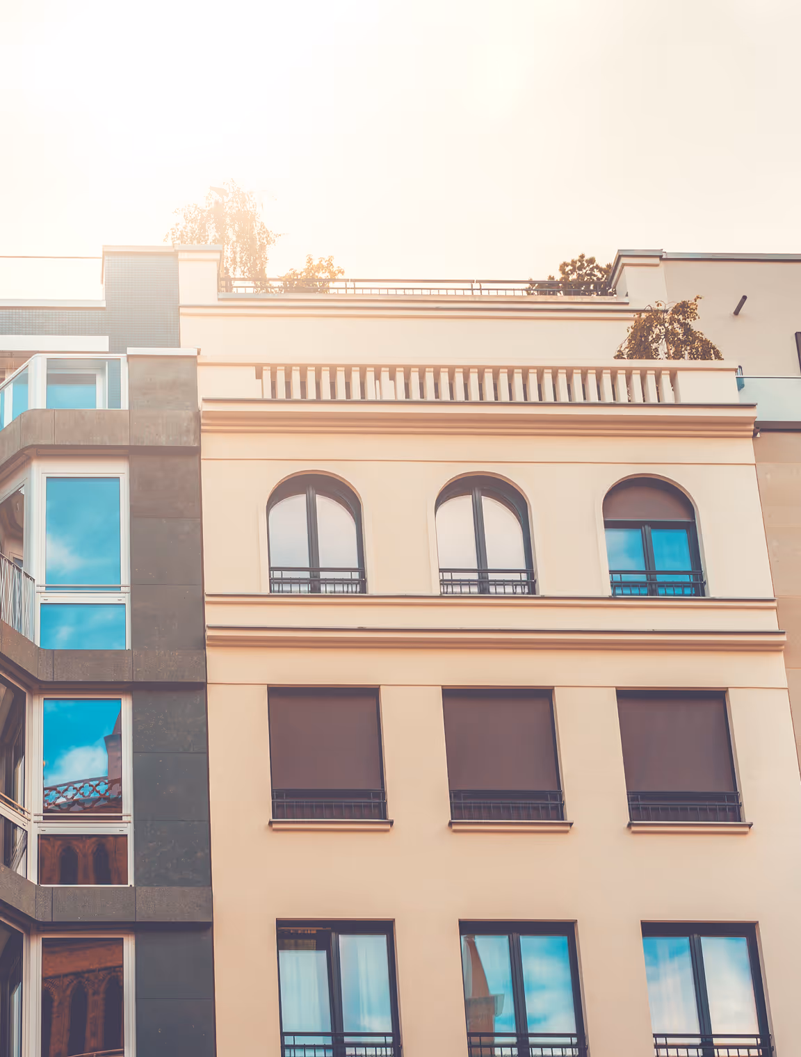 Fassade eines mehrstöckigen Gebäudes mit großen Fenstern, einige sind mit Rollläden bedeckt, unter hellem Sonnenlicht.