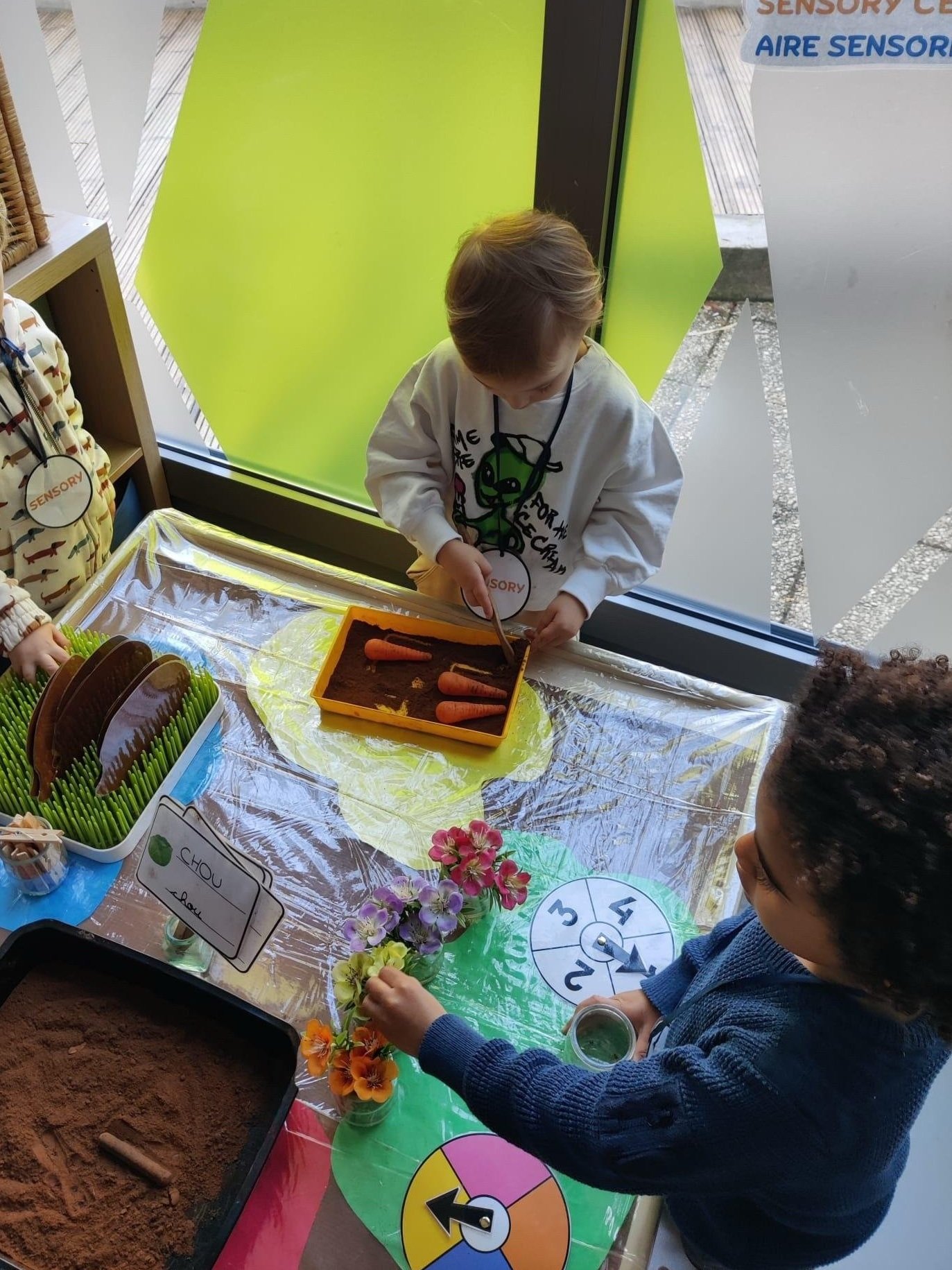Enfants en activité sensorielle en très petite section de maternelle bilingue – découverte des légumes, fleurs et textures dans un atelier d'éveil à l'École M
