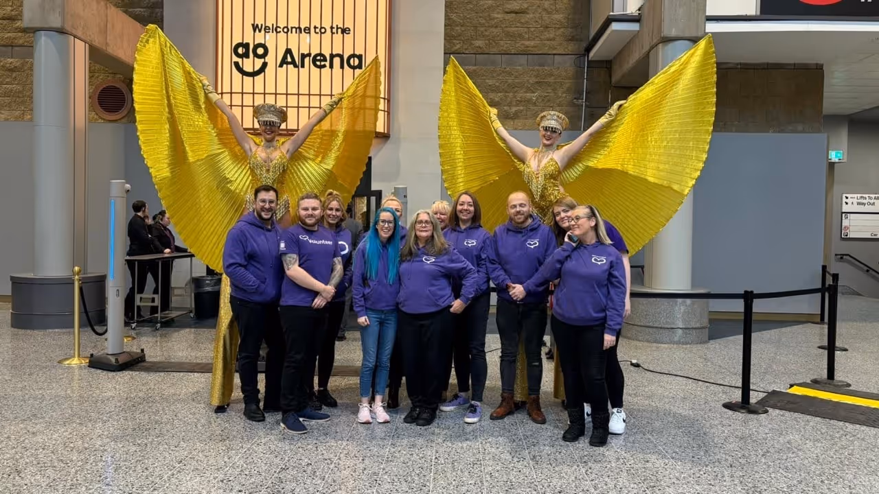 Group of people wearing purple hoodies standing in front of two performers with large golden wings inside a building with a sign that reads 'Welcome to the Arena'.