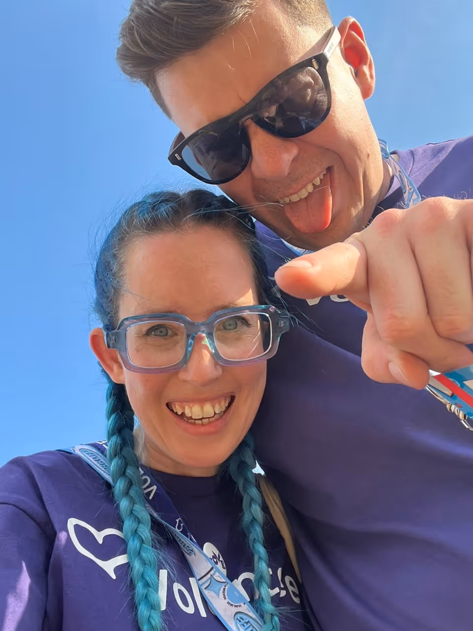 Smiling woman with blue braided hair and clear glasses and man wearing dark sunglasses sticking out his tongue and pointing at the camera, both in purple shirts under a clear blue sky.