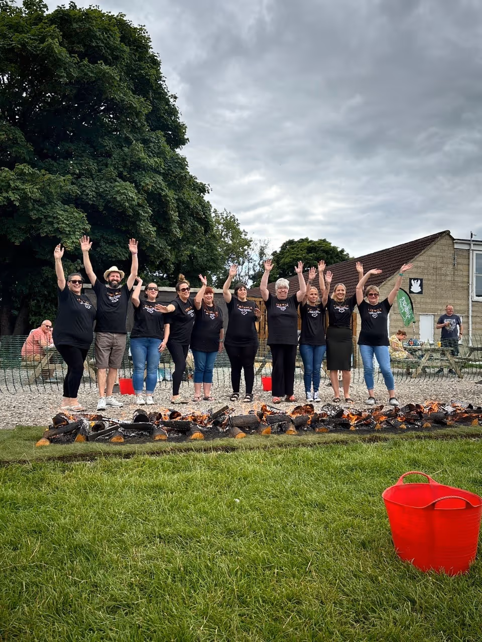 Group of people standing behind a line of burning logs with hands raised, outdoors on grass under a cloudy sky.