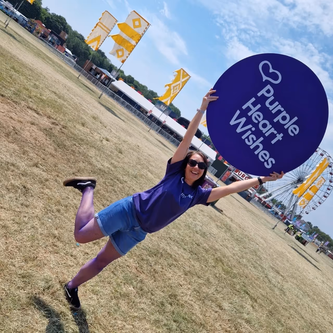 Smiling woman wearing purple tights and a Purple Heart Wishes shirt holding a large purple sign at an outdoor festival with yellow flags and a Ferris wheel in the background.