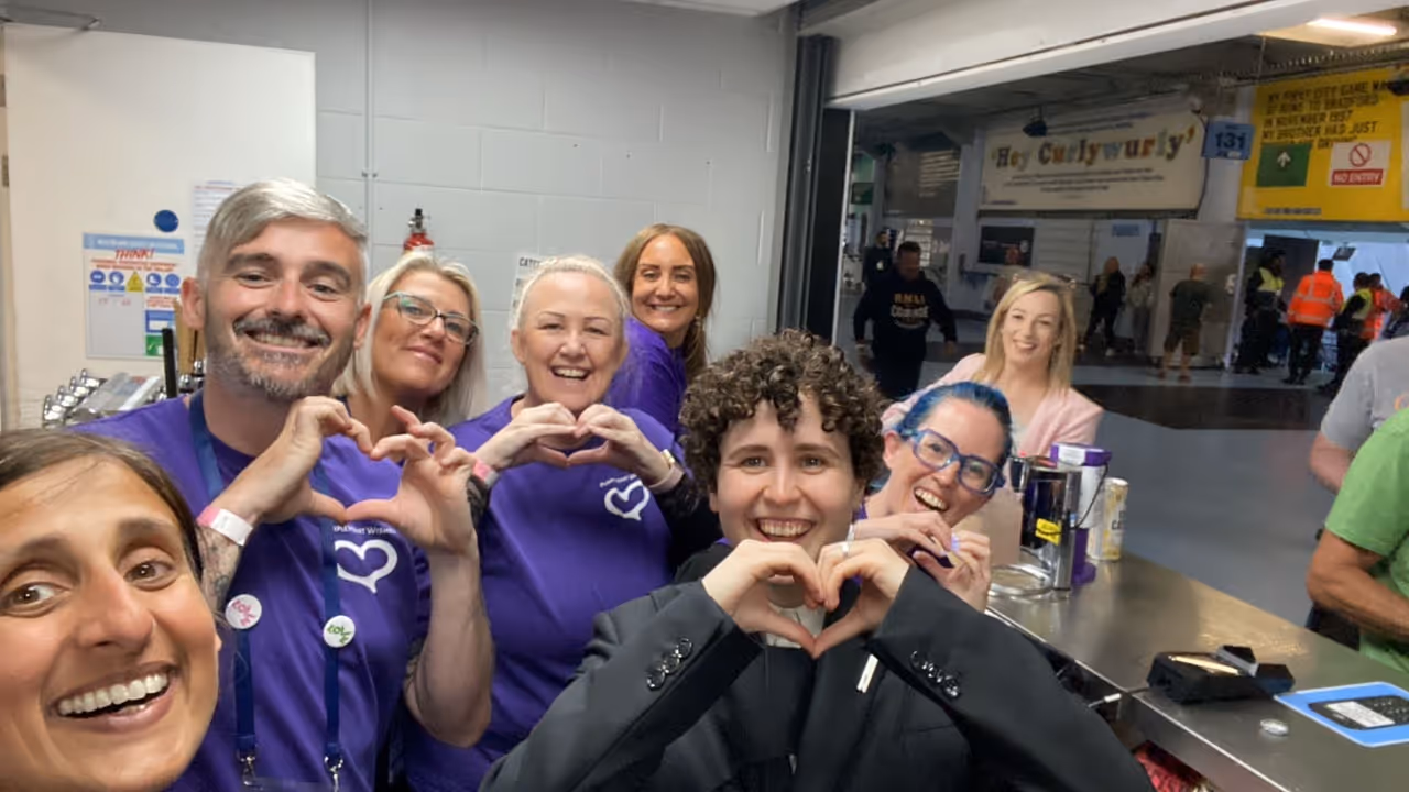 Group of smiling people indoors, several wearing purple shirts making heart shapes with their hands.