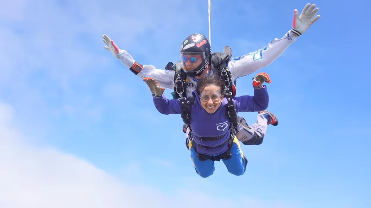 Two people tandem skydiving in freefall against a clear blue sky, one smiling at the camera.