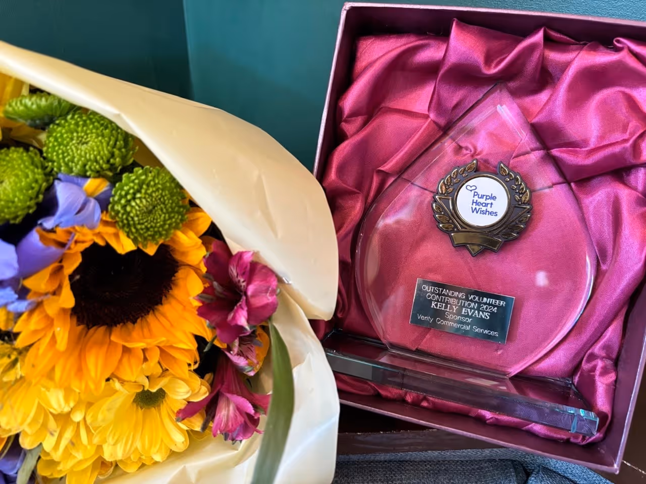 Award in a red satin-lined box labeled 'Outstanding Volunteer Contribution 2024 Kelly Evans' next to a colorful bouquet of flowers including sunflowers and green chrysanthemums.