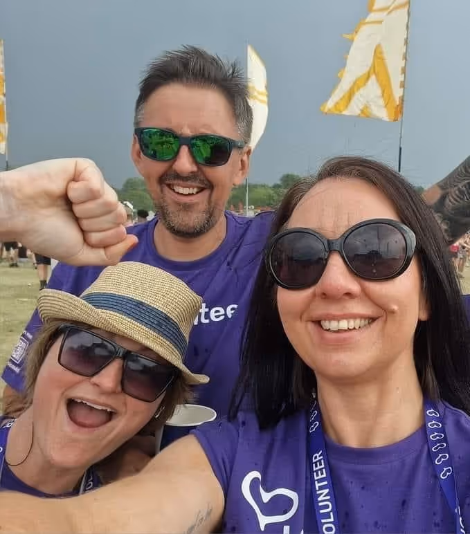 Three smiling volunteers wearing sunglasses and purple shirts posing together outdoors at a sunny event with flags in the background.
