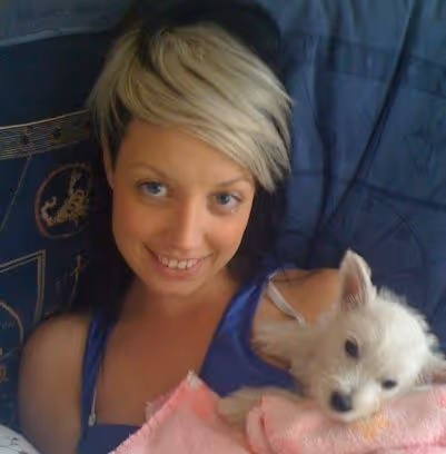 Smiling woman with short blonde and black hair holding a small white dog wrapped in a pink blanket.