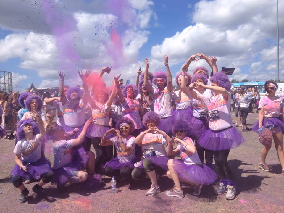 Group of people wearing purple tutus and wigs celebrating with purple and pink powder in the air under a blue sky with clouds.