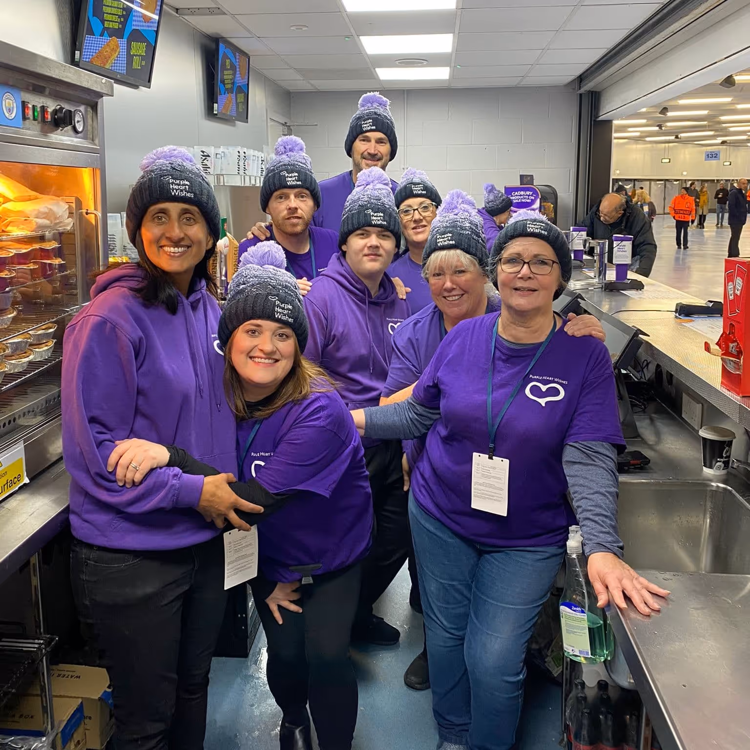 Group of nine people wearing purple shirts and matching knit hats with pom-poms, smiling inside a food service area.