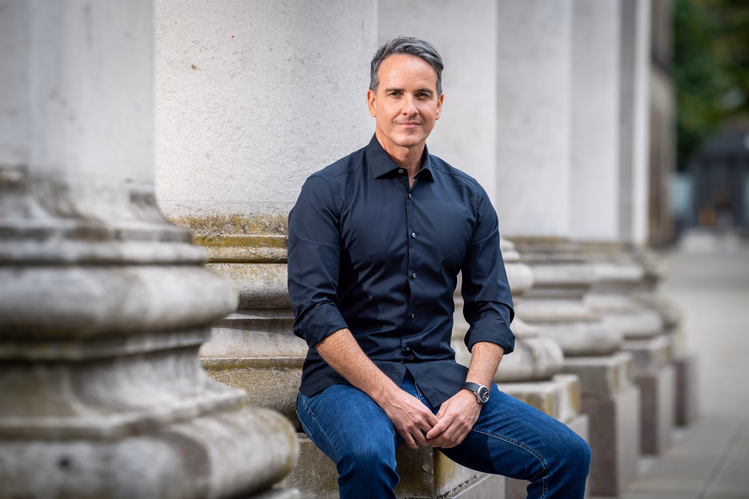 Man with gray hair wearing a black button-up shirt and blue jeans sitting on a stone ledge among large white columns.