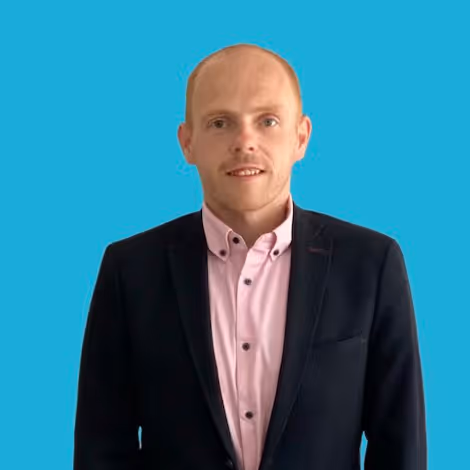 Man with light skin and short hair wearing a black blazer over a pink button-up shirt against a blue background.