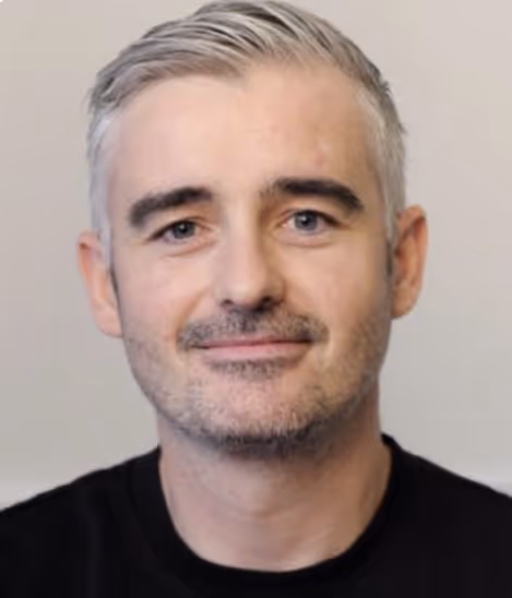 Smiling middle-aged man with short gray hair and light facial stubble wearing a black shirt against a plain background.