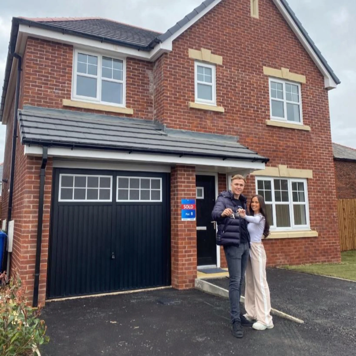 Smiling couple standing in front of a newly purchased two-story brick house holding keys with a sold sign on the wall.