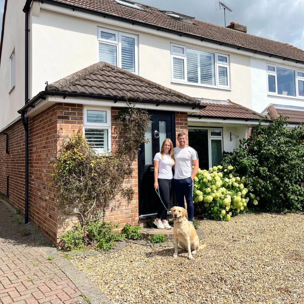 Smiling couple in casual clothes standing with their labrador dog on a gravel driveway in front of a suburban brick house with white upper walls and flower bushes.