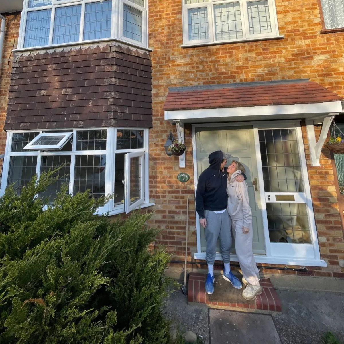 Couple standing on the doorstep of a brick house, embracing and looking at each other warmly.
