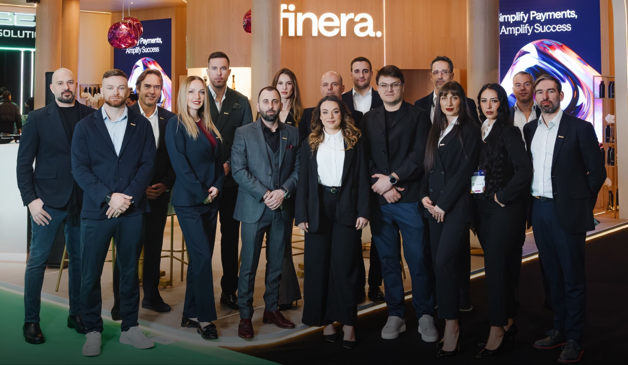A professional group portrait of the Finera team standing together at their exhibition booth during the ICE Barcelona event, featuring modern branding and "Simplify Payments, Amplify Success" digital displays in the background.
