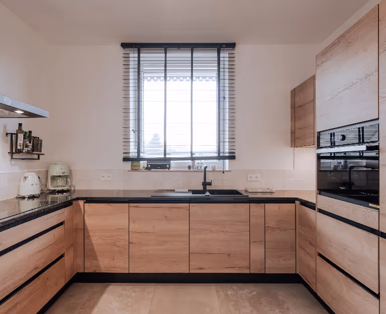 Modern kitchen with light wood cabinets, black countertop, window with blinds above sink, and built-in oven.