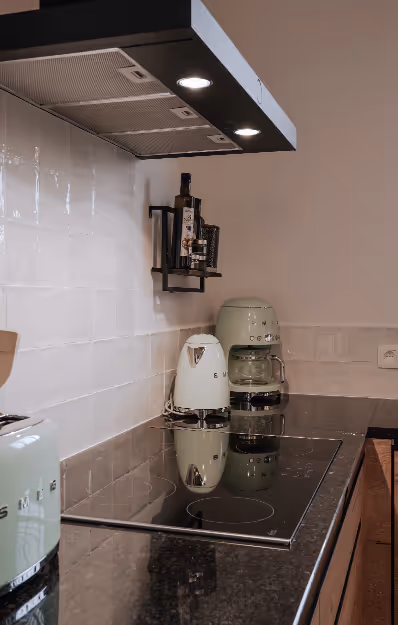 Modern kitchen countertop with black glass stovetop, vintage-style SMEG appliances including a kettle, coffee maker, and toaster, and a small wall shelf holding olive oil and spices.