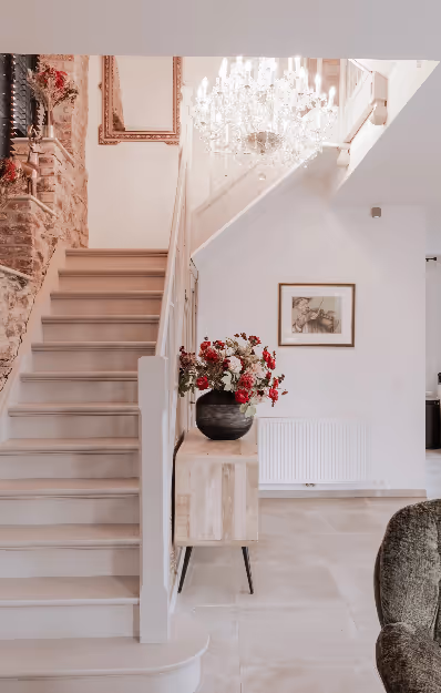 Interior view of a modern home with a white staircase, wooden side table holding a vase with red and white flowers, a framed picture on the wall, and a crystal chandelier overhead.