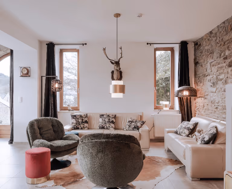 Cozy living room with cream sofas, floral cushions, two green armchairs, a mounted deer head, and exposed stone wall.