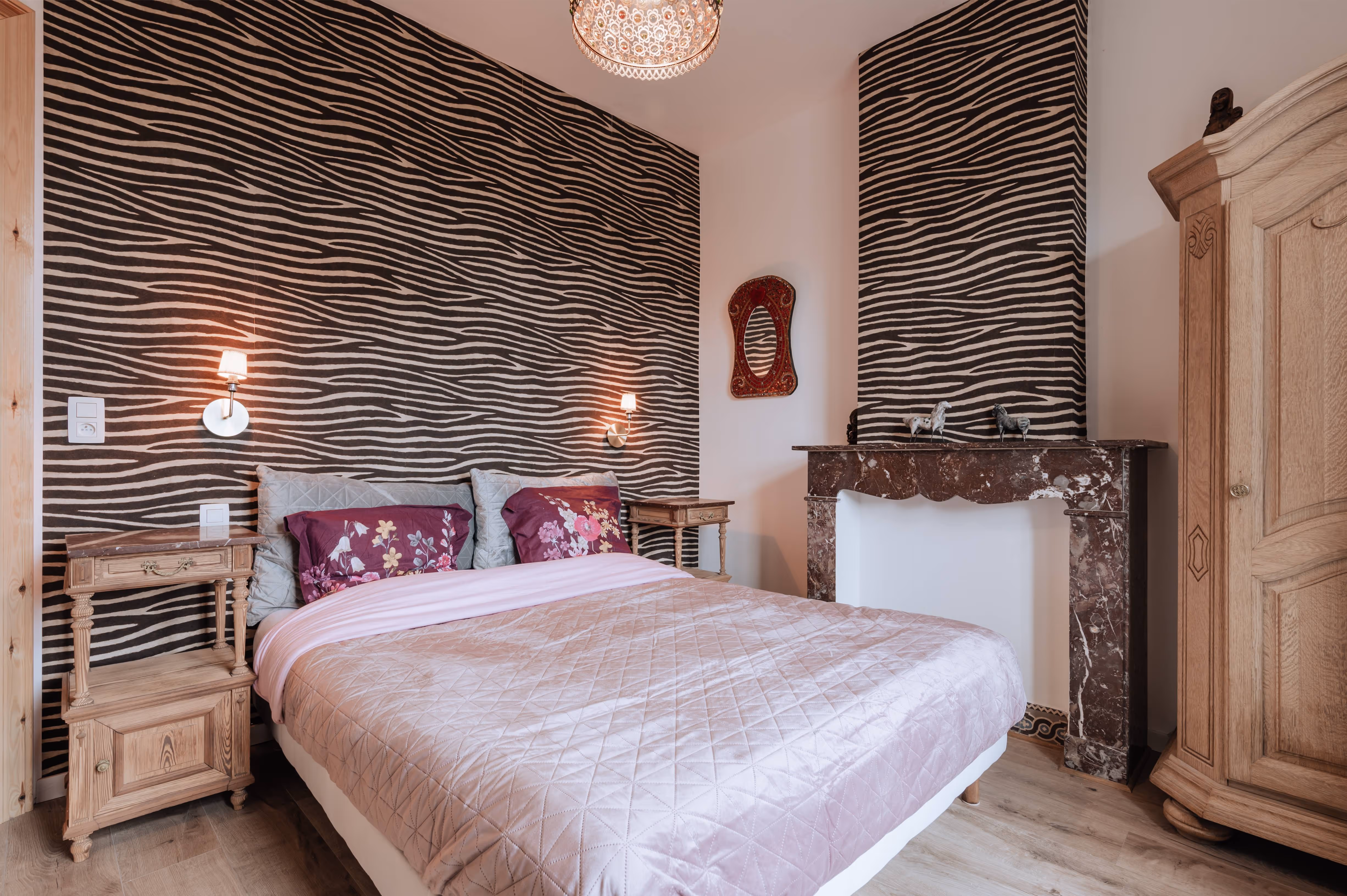 Bedroom with a double bed featuring lavender quilt and floral pillows, zebra-patterned accent wall, wooden nightstands, decorative fireplace, and wooden wardrobe.