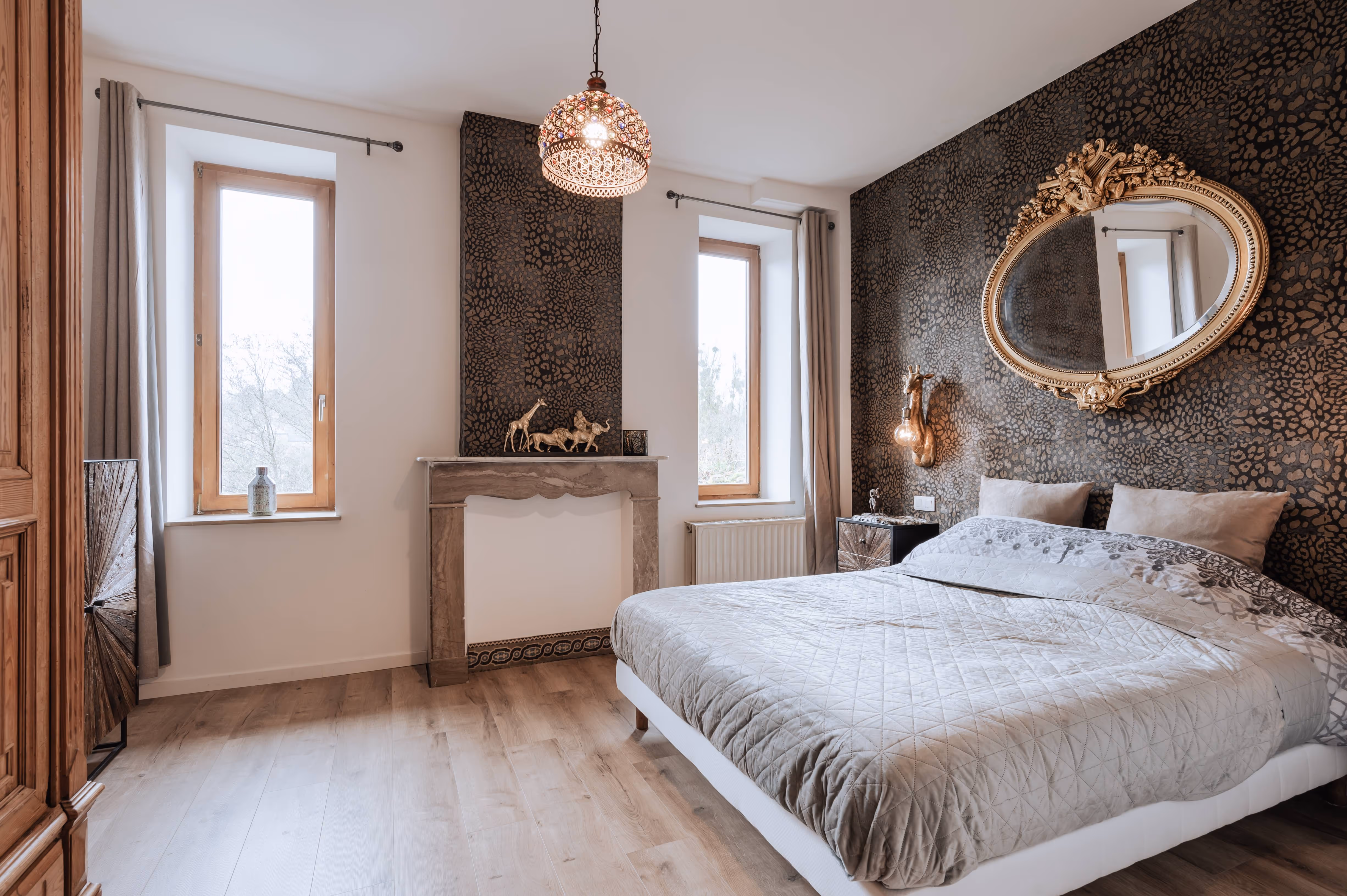 Modern bedroom with patterned dark accent walls, large bed with gray bedding, ornate oval mirror, decorative giraffe lamp, and two tall windows with gray curtains.